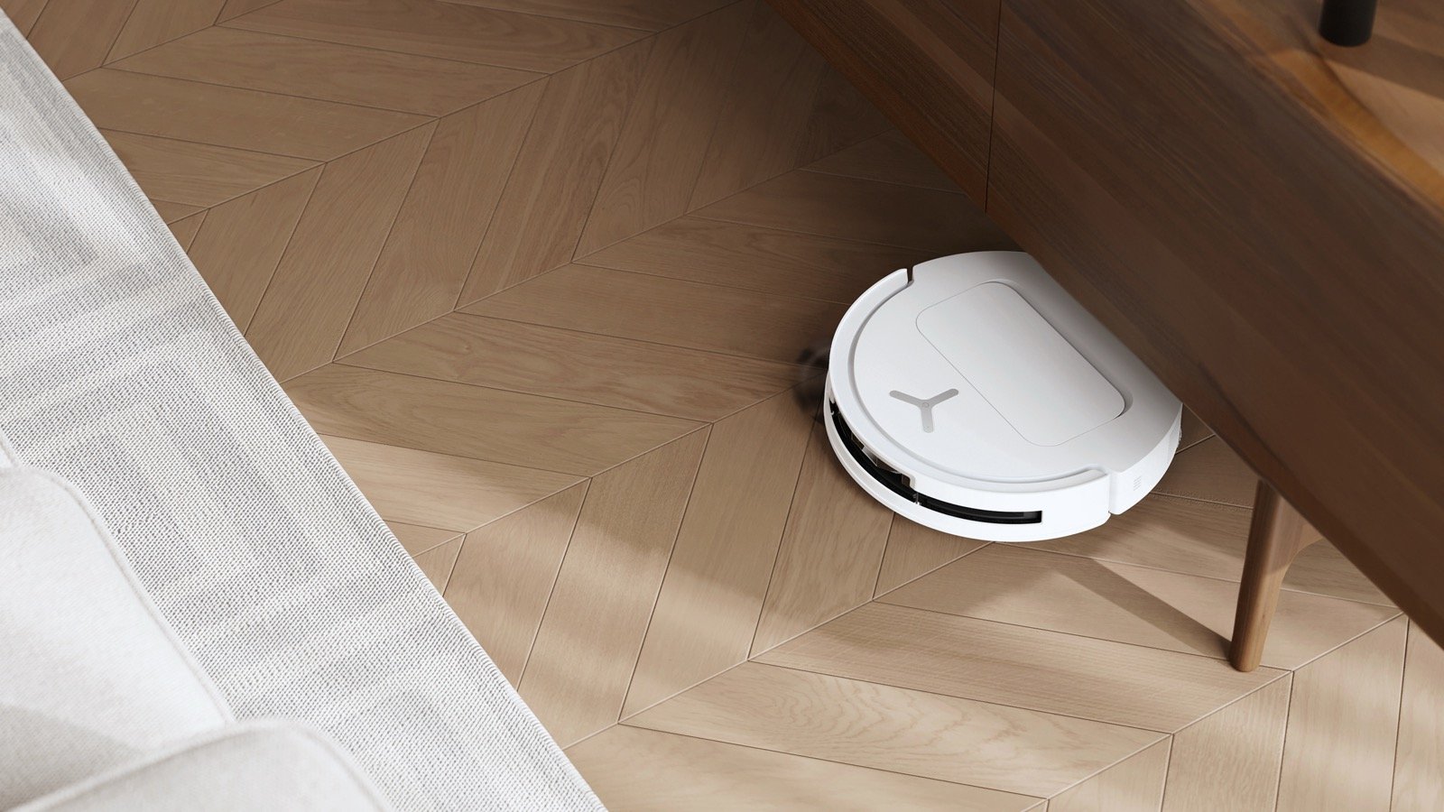 White robot vacuum on herringbone wood flooring as it passes under a wooden table, manufacturer still, no people